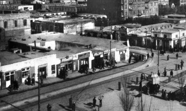 Schwarz-weiß-Foto einer Stadtstraße mit Fußgängern, umgeben von Gebäuden, Bäumen, Pfählen und Drähten.
