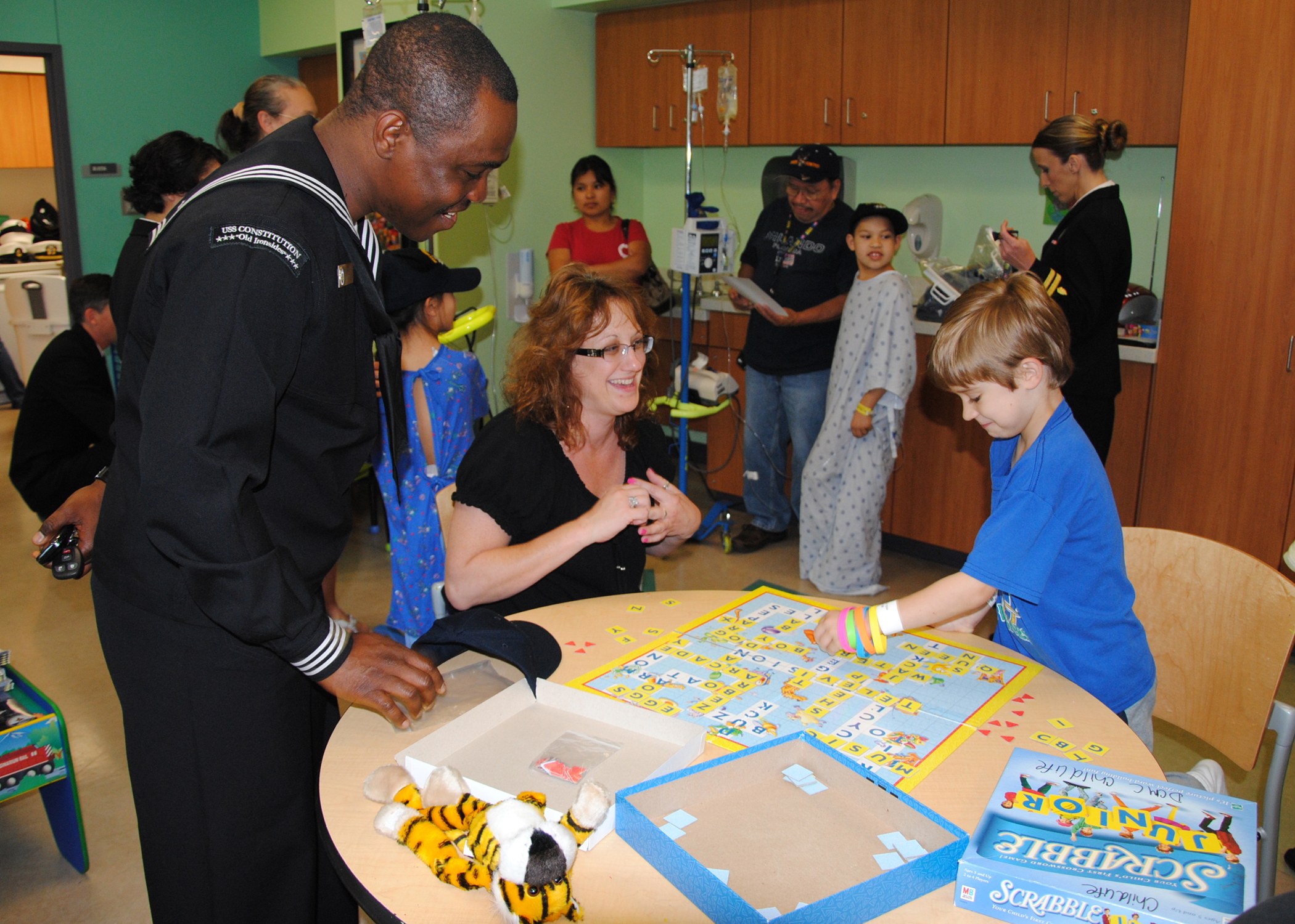 Gruppe von Menschen, die um einen Tisch versammelt sind und ein Brettspiel mit Spielzeugen, Boxen und anderen Gegenständen darauf spielen, in einem Raum mit Schränken, einer Salzlösung auf einem Ständer, einer Tür und einer Wand im Hintergrund.