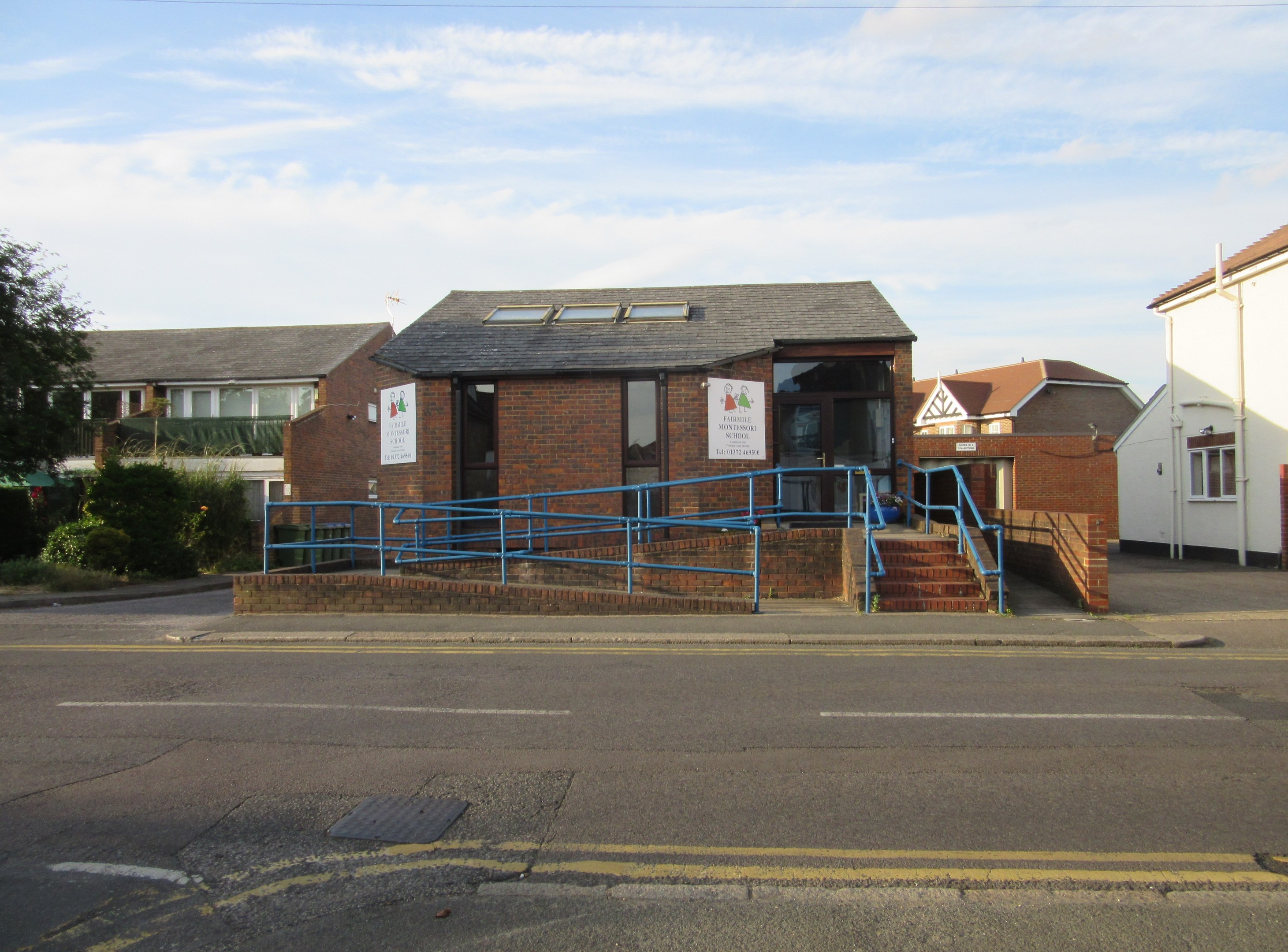 Ein Schulgebäude mit blauer Begrenzung, umgeben von Häusern, Treppen, Geländern, Texttafeln, Pflanzen, Bäumen und einem bewölkten Himmel.