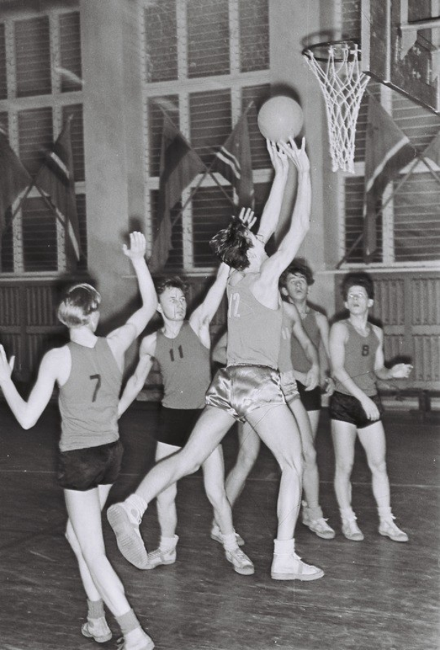 Schwarz-weiß-Foto von jungen Männern, die Basketball in einer Turnhalle spielen, mit einem Basketballkorb in der oberen rechten Ecke und Flaggen im Hintergrund.