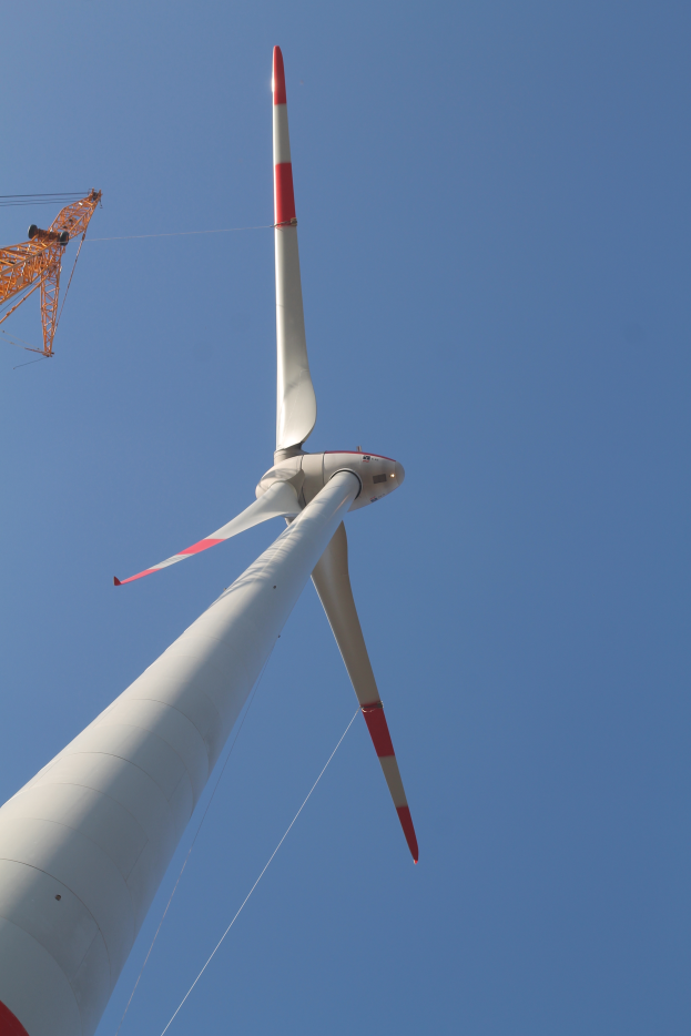 Eine weiße und rote Windkraftanlage mit einem Kran im Hintergrund vor einem klaren blauen Himmel.