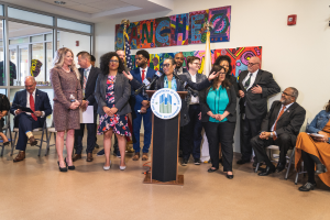 Eine Gruppe von Menschen, die vor einem Podium mit Mikrofon und einer Tafel mit der Aufschrift "Sancho Stadtrat joins City Council" stehen, mit Flaggen, Feuerlöscher und Deckenleuchten an der Rückwand.