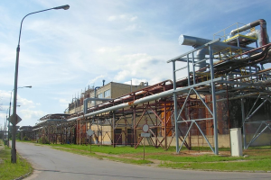 Große Fabrik mit ausgedehnter Rohrleitung, umgeben von Gebäuden, Straßeninfrastruktur, Grünflächen und einem bewölkten Himmel.