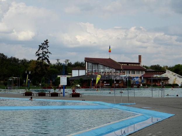Ein großes Schwimmbecken voller Menschen, umgeben von Pfosten, Bänken, Topfpflanzen, einem Schild, einem Fahnenmast mit Flagge, einem Gebäude mit Fenstern, Straßenlaternen, einer Baumgruppe, unter einem bewölkten Himmel.
