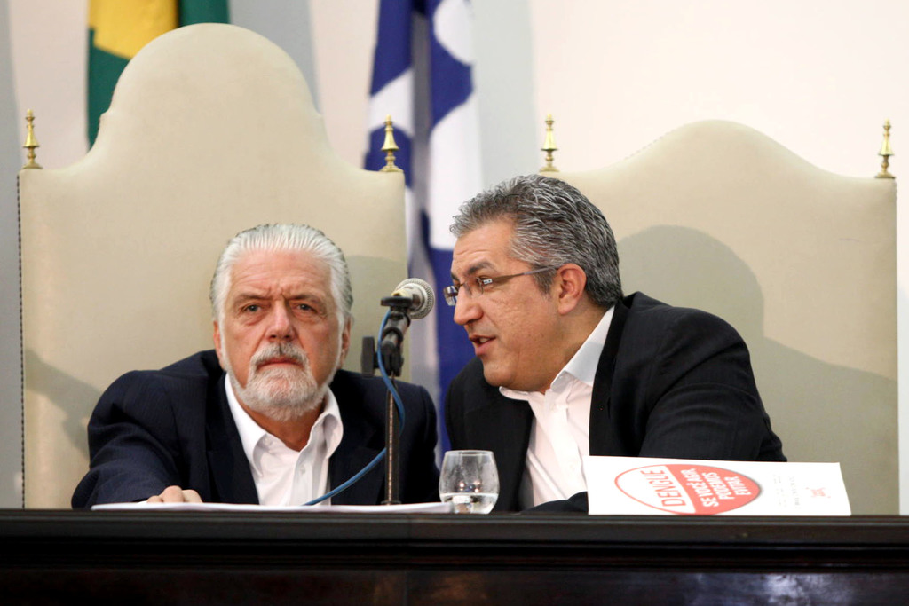 Zwei Männer sitzen an einem Tisch mit einem Mikrofon davor, mit Flaggen oben im Bild.