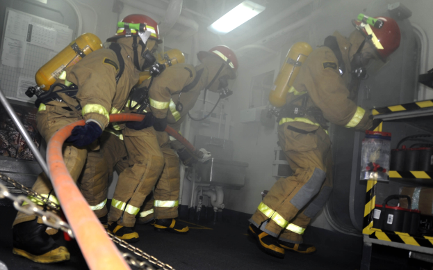 Feuerwehrleute in Schutzausrüstung arbeiten an einem Feuerwehrauto mit Ausrüstungsgestellen und Hintergrundinfrastruktur.