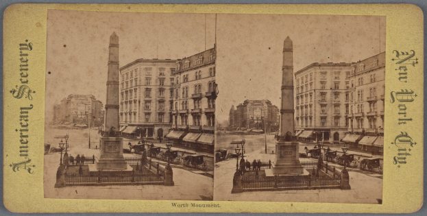 Altes Schwarz-Weiß-Foto einer Stadtstraße mit einem zentralen Denkmal, umgeben von Gebäuden, Straßenmöbeln, geparkten und fahrenden Fahrzeugen, Fußgängern auf dem Gehweg und einem sichtbaren Himmel.