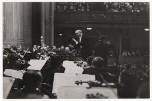 Ein Schwarz-Weiß-Foto eines Dirigenten mit einem Dirigentenstab, der vor einem Orchester mit verschiedenen Instrumenten steht, mit Notenpulten und einem Geländer im Hintergrund.