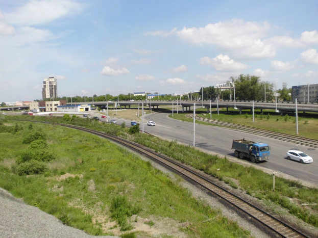 Eine Eisenbahnschiene verläuft durch eine Stadtlandschaft mit Fahrzeugen, Grünflächen, Polen, Brücken, Gebäuden und einem bewölkten Himmel im Hintergrund.