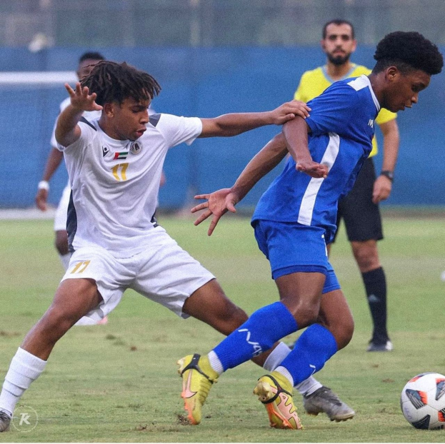 Zwei Männer spielen Fußball auf einem Rasenfeld mit einem Zaun im Hintergrund, einer scheint einen Sieg zu feiern.
