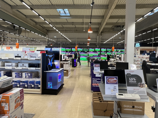 Ein großer Raum in einem Walmart-Laden voller Elektronik wie Computer und Drucker, die in Regalen angeordnet sind, mit Menschen auf dem Boden, einer Säule, Schildern, Deckenlampen und Ventilatoren.
