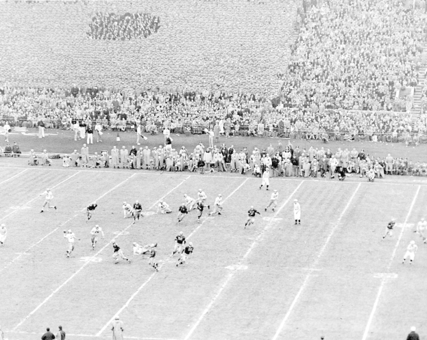 Schwarzes Foto eines Fußballspiels im Gange, mit Spielern auf dem Feld und einer Zuschauermenge im Hintergrund.