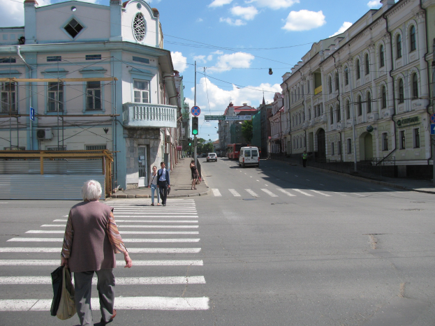 Alte Frau überquert die Straße vor einem Gebäude, umgeben von Fußgängern, Fahrzeugen, Schildern, Strommasten, Bäumen und einem bewölkten Himmel.
