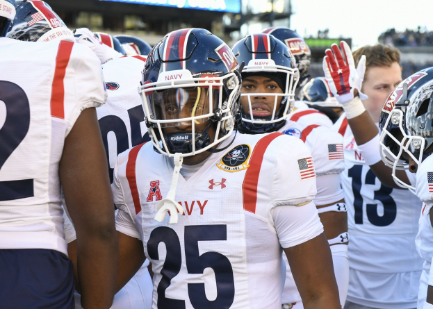 Gruppe von Männern in Fußballausrüstung auf einem Feld mit Helmen und Handschuhen, Hintergrund enthält unscharfe Zuschauer, ein Scoreboard mit "Auburn vs Virginia Tech - 11/28/15 College Football Pick, Odds, and Prediction" und Himmel.