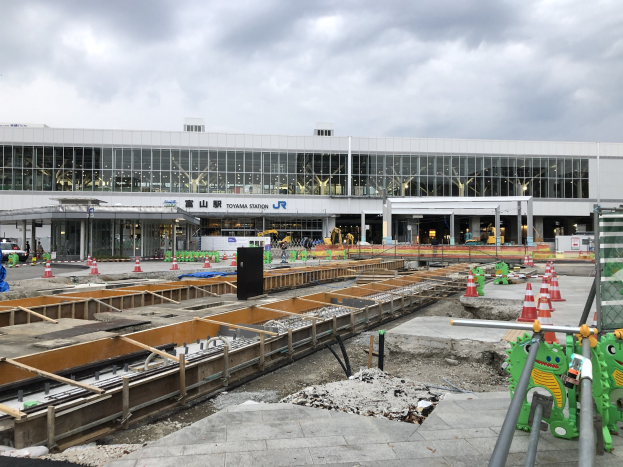 Baustelle am Toyama-Bahnhof mit einem großen Gebäude im Hintergrund, umgeben von Fahrzeugen, Absperrungen, Pfählen, Schildern und anderen Gegenständen mit einer bewölkten Himmel.