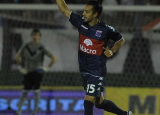 Ein Fußballer auf dem Boden, der einen Torerfolg feiert, mit einer unscharfen Menge und einer weiteren Person im Hintergrund hinter einer Tafel mit Text.