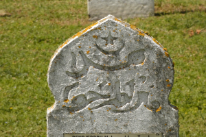 Ein steinerner Grabstein mit arabischer Schrift in einem grünen Friedhof.