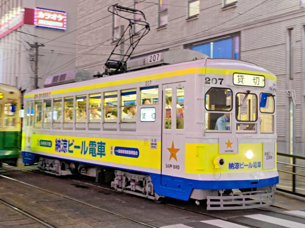 Eine gelbe und blaue Straßenbahn auf einer Stadtstraße bei Nacht mit Menschen drinnen, Gebäude mit Fenstern, Strommasten mit Kabeln und Schilder mit Text im Hintergrund.