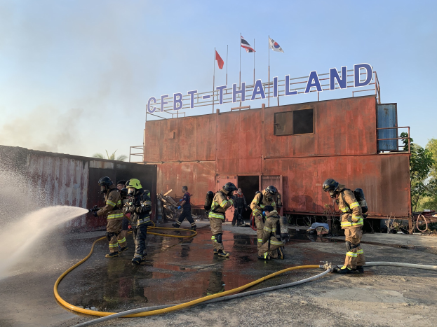 Feuerwehrleute in Helmen sprühen Wasser auf ein Gebäude mit Text, während Fahnen, Bäume und ein klarer Himmel im Hintergrund zu sehen sind.
