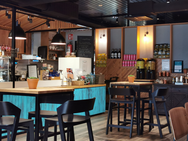 Innenraum eines Restaurants mit Holztischen, Stühlen, einer Topfpflanze auf einem Tisch, einer Schiefertafelmenü, einem Kühlschrank, einem Feuerlöscher, Regalen mit Flaschen, Schränken mit Gegenständen, einem Rahmenfoto an der Wand und Deckenleuchten.