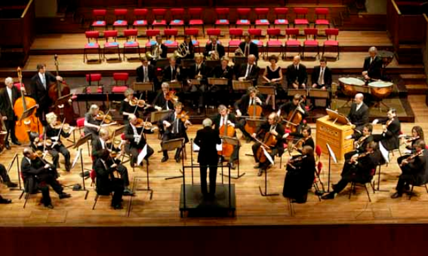 Ein großes Orchester auf einer Bühne mit einem Dirigenten vorne, der verschiedene Instrumente spielt, mit Stühlen, Ständern und Treppen im Hintergrund.