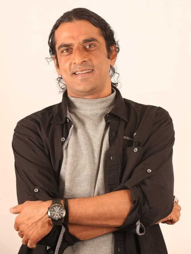 Ein Mann in einem schwarzen und aschfarbenen Kleid mit überschlagenen Armen steht vor einem wei├čen Hintergrund und tr├Ągt eine Uhr, scheinbar in Gedanken versunken.