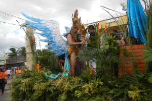 Eine Gruppe von Menschen geht neben einem blumen- und pflanzengeschmückten Faschingswagen mit kostümierten Personen die Straße entlang.