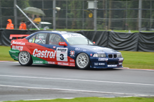 Ein Auto rennt auf einer Rennstrecke mit Gras auf beiden Seiten, mit Menschen in orangefarbenen Jacken und Schirmen hinter einem Maschendrahtzaun mit schwarzem Stoff und Bäumen im Hintergrund.