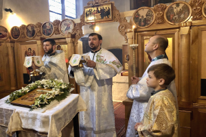 Eine Gruppe von Menschen, die um einen Tisch in einer Kirche stehen, Bücher halten, mit einem Tuch und einem Blumenstrauß auf dem Tisch und Fotorahmen, Lampen, einem Fenster und einem hölzernen Objekt im Hintergrund.