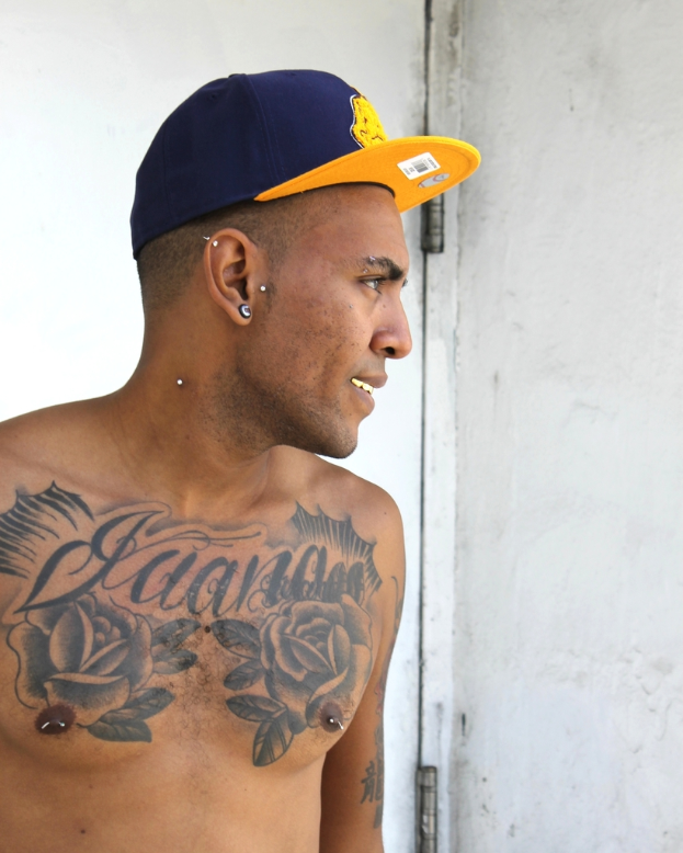Eine Person mit einer Mütze mit einem Aufkleber mit einem Barcode und Bildern, mit sichtbaren Tattoos am Körper, die vor einer Tür und einer Wand steht.