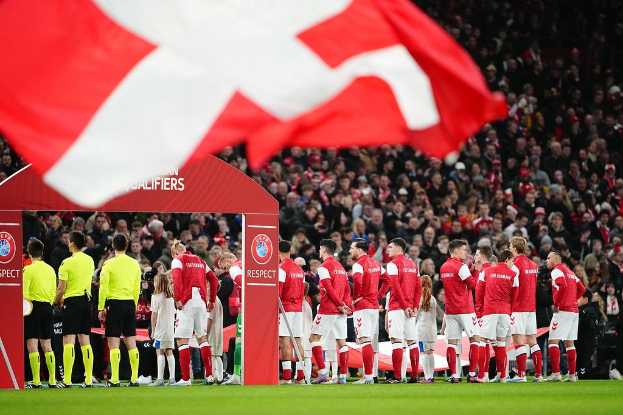 Eine Gruppe von Menschen auf einem Fußballfeld mit einer roten und weißen Flagge im Vordergrund, einem Bogen mit "Bayern Munich vs Bayern Munich Betting Tips & Preview" im Hintergrund und einer großen Menge im Stadion.