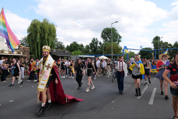 Eine Gruppe von Menschen marschiert bei der Christopher Street Day Parade 2018 mit einer Regenbogenflagge und Musikinstrumenten, einige tragen Mützen, vor einer Kulisse aus Laternenmasten, Bäumen, Hütten und einem bewölkten Himmel.
