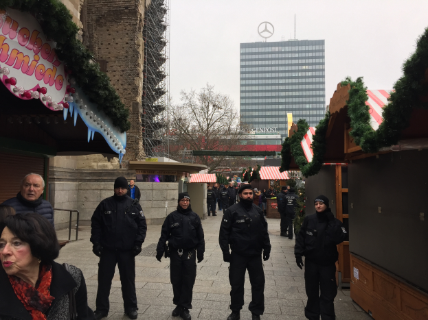 Eine Gruppe von Polizisten in schwarzen Uniformen steht vor einem festlich geschmückten Weihnachtsmarkt in Berlin, Deutschland, mit Gebäuden, Bäumen und einem klaren blauen Himmel im Hintergrund und einer Tafel mit Schrift auf der linken Seite des Bildes.