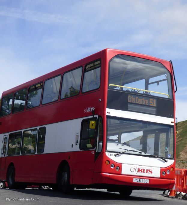 Ein roter Doppeldeckerbus mit der Aufschrift 'Stadtbus' fährt auf einer Straße mit einem Verkehrskegel im Vordergrund und einem Hügel im Hintergrund.