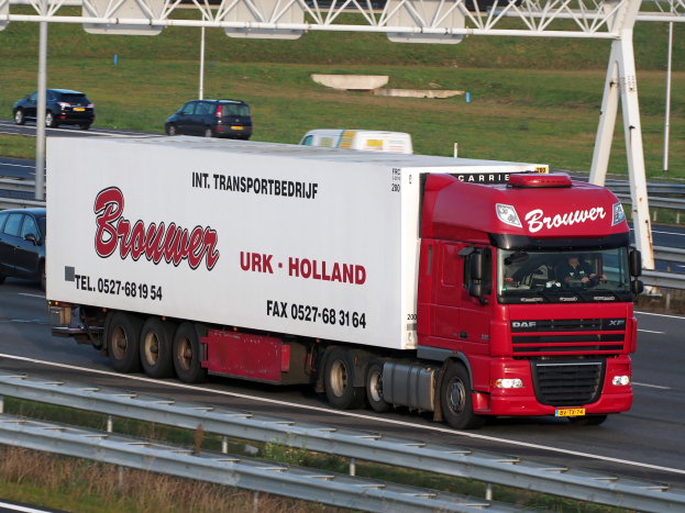 Ein roter und weißer Lastwagen mit Text darauf fährt auf einer Autobahn, umgeben von anderen Fahrzeugen, mit einer Leitplanke am Straßenrand, Gras im Hintergrund und einem Tor mit Pfosten.