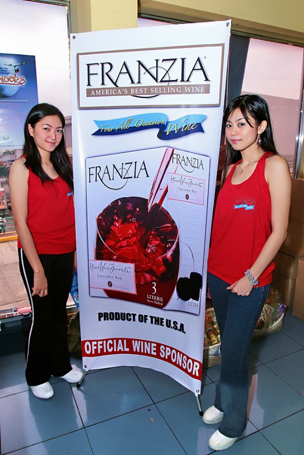 Zwei Frauen neben einem Plakat für Franzia, Americas bestverkaufenden Wein, mit einem Glasfenster im Hintergrund.