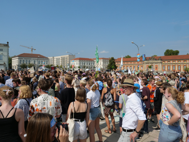 Eine große Menschenmenge steht vor einem Gebäude mit Fenstern, Laternenmasten, Fahnenmasten und Bäumen unter einem bewölkten Himmel, einige halten Kameras während einer Demonstration.