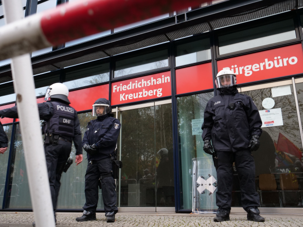 Polizeibeamte in Uniform vor einem Glasfrontgebäude mit Helm und Handschuhen, links ein Mast und Schilder mit der Aufschrift "Friedrichshain Kreuzberg" im Hintergrund.
