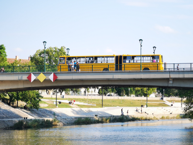 Ein gelber Schulbus überquert eine Brücke über einen Fluss, mit Menschen, die über die Brücke gehen und stehen, umgeben von saftigem Grün und Bäumen, mit Gebäuden und einem klaren blauen Himmel im Hintergrund.