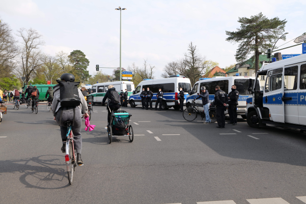 Eine Gruppe von Menschen, die Fahrräder auf einer Straße fahren, die von Polizeiwagen und anderen Fahrzeugen gesäumt ist, einige tragen Helme und Taschen, andere schieben Kinderwagen, mit Bäumen, Laternenmasten, Verkehrsampeln, Schildern, Gebäuden und einem bewölkten Himmel im Hintergrund.