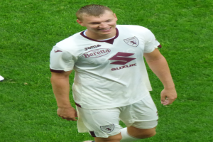 Ein Mann in einem weißen Hemd und Shorts lächelt, während er auf einem Fußballfeld geht und Schuhe trägt.