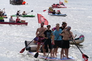 Eine Gruppe von Menschen auf Paddle-Boards im Wasser, einige halten Paddel, mit Booten und Flaggen im Hintergrund bei der 2016er Stand-Up-Paddleboard-Festival.