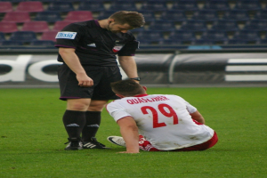 Ein Fußballspieler in Sportkleidung sitzt neben einem Schiedsrichter auf dem Boden, beide in einer Stadionumgebung mit Tafeln und Stühlen im Hintergrund.