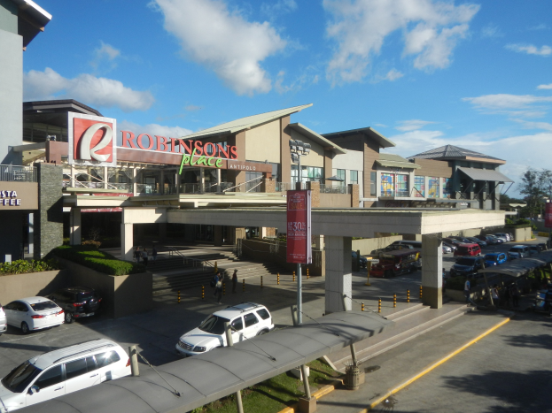 Ein belebtes Einkaufszentrum mit Gebäuden, Schaufenstern, Fahrzeugen, Fußgängern, Straßenmöbeln, Grünflächen und einem bewölkten Himmel.