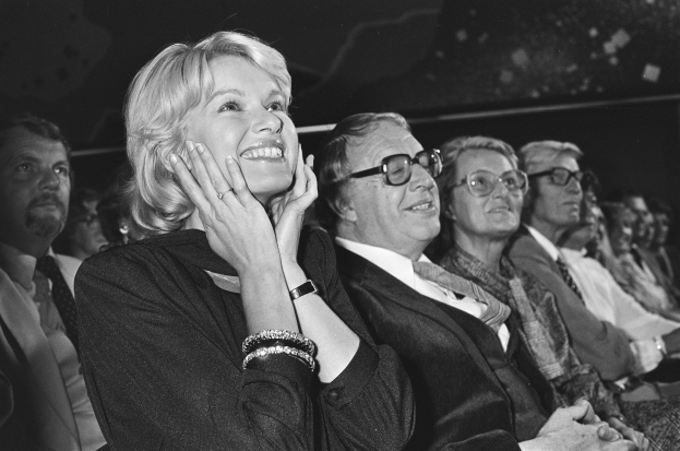 Eine Gruppe von Menschen, darunter eine Schwiegermutter mit gefalteten Händen, sitzt zusammen vor einem Publikum, alle tragen Brillen und lächeln, auf einem Schwarz-Weiß-Bild.