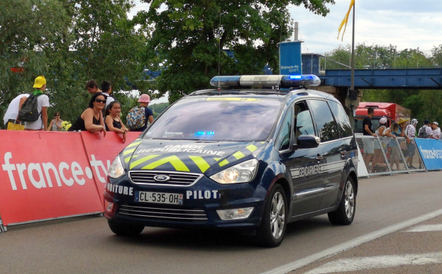 Polizeiauto fährt an einer Menge vorbei, die sich an einem Geländer mit Schildern befindet, daneben Bäume, eine Brücke, eine Fahne und einen bewölkten Himmel im Hintergrund.