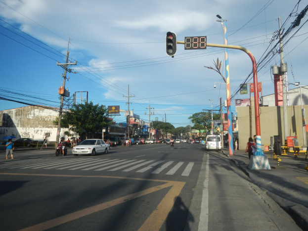 Eine Straßenkreuzung in einer Stadt mit einer Ampel, Fahrzeugen, Fußgängern, Strommasten, Bäumen, Gebäuden und Verkehrszeichen unter einem bewölkten Himmel.