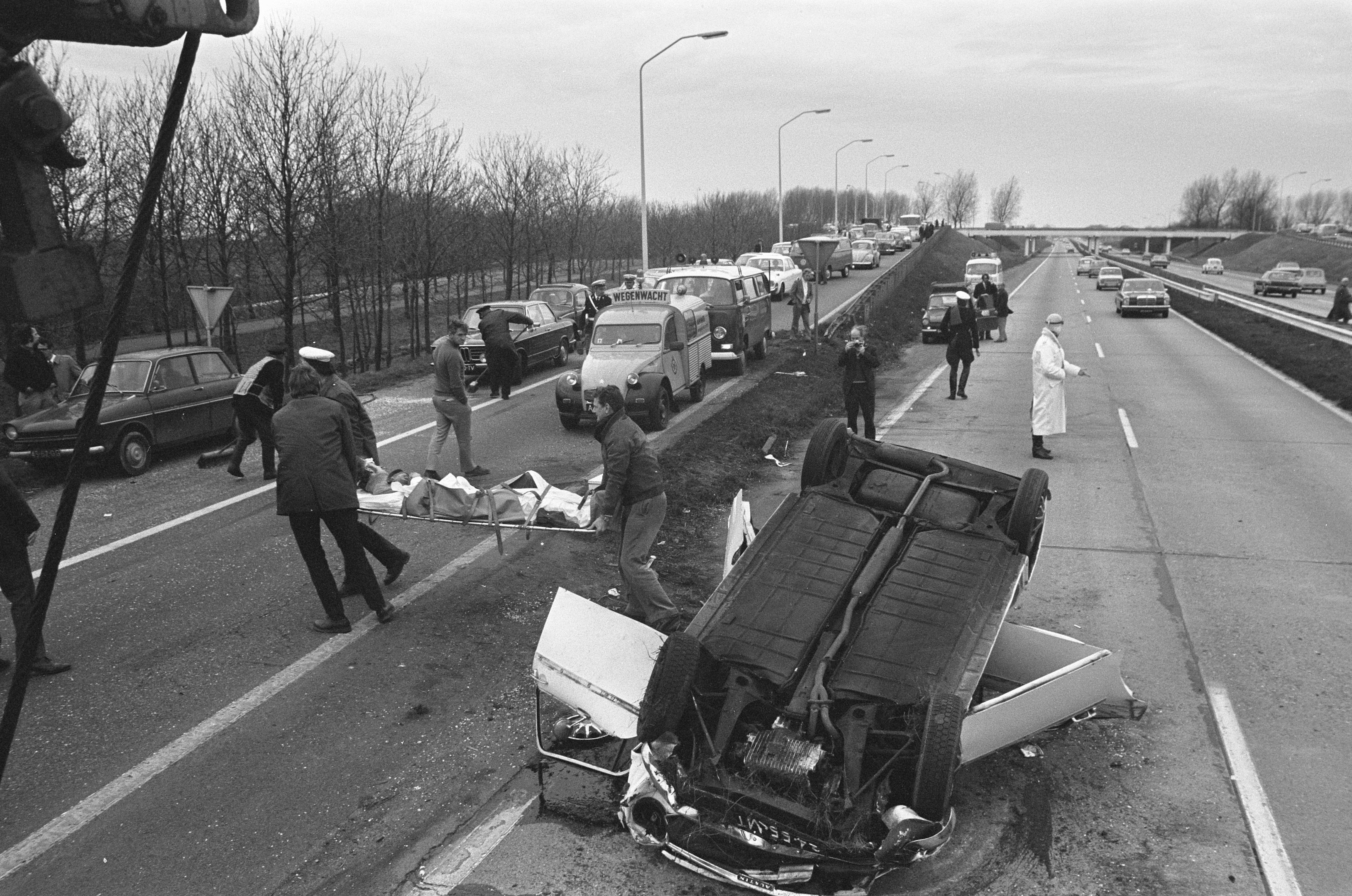 Ein Schwarz-Weiß-Foto eines umgestürzten Autos am Straßenrand mit beschädigten Fahrzeugen in der Nähe und einer Gruppe von Menschen drumherum, vor einem Hintergrund aus Laternenmasten, Bäumen, einer Brücke und dem Himmel.
