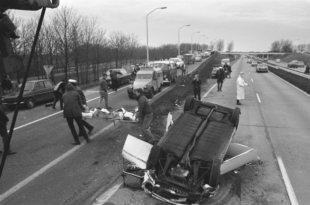 Ein Schwarz-Weiß-Foto eines umgestürzten Autos am Straßenrand mit beschädigten Fahrzeugen in der Nähe und einer Gruppe von Menschen drumherum, vor einem Hintergrund aus Laternenmasten, Bäumen, einer Brücke und dem Himmel.