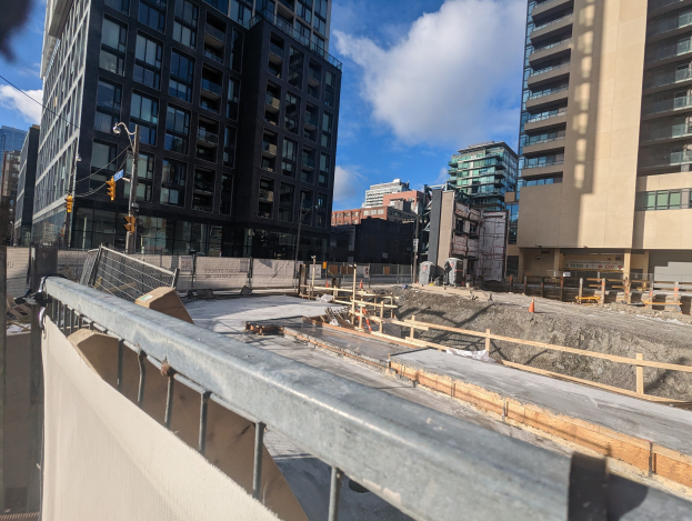 Baustelle in Toronto, Ontario mit einem großen Bauwerk im Bau, umgeben von Absperrungen, Pfählen, Verkehrszeichen und Schildern unter einem bewölkten Himmel.